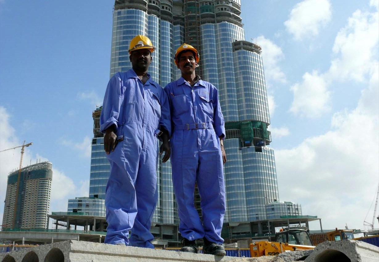 Construction workers are silhouetted against a skyscraper, illustrating migration and labor in the Gulf region.